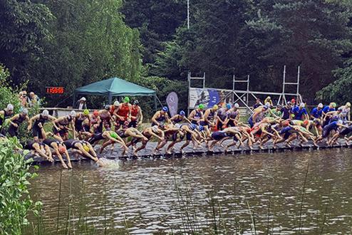 Sportler springen vom Steg ins Wasser bei einem Triathlon-Event in einem bewaldeten Umfeld. Grüne Vegetation umgibt den See.
