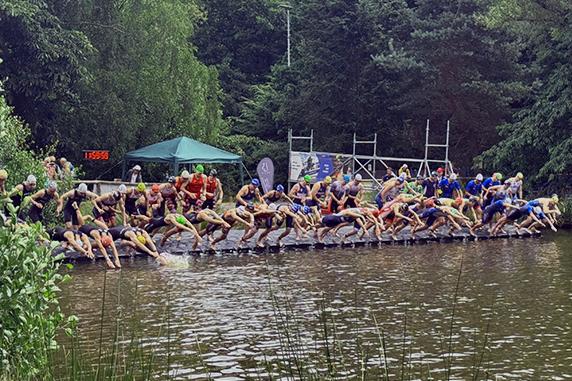 Sportler springen vom Steg ins Wasser bei einem Triathlon-Event in einem bewaldeten Umfeld. Grüne Vegetation umgibt den See.