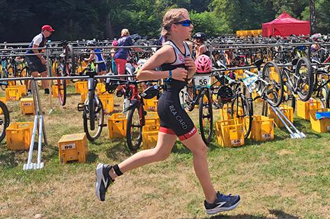 Junge Sportlerin in Wettkleidung läuft durch eine Transitionzone mit Fahrrädern und gelben Transportboxen.