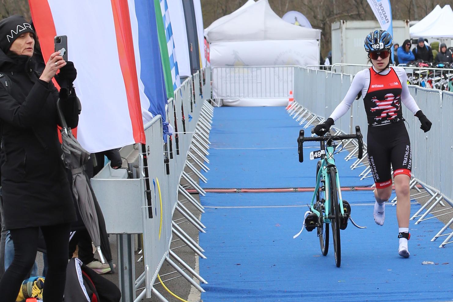 Athlet in Triathlonsportkleidung schiebt ein Rennrad auf einer blauen Fläche, während Zuschauer in der Umgebung stehen.