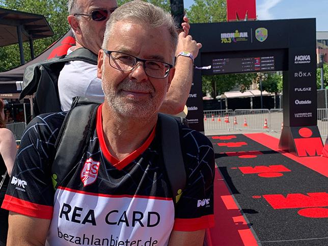 Mann mit Brille und rot-weißem Sportshirt steht an einer Absperrung vor dem Ironman-Event, Hintergrund mit Eventbanner.