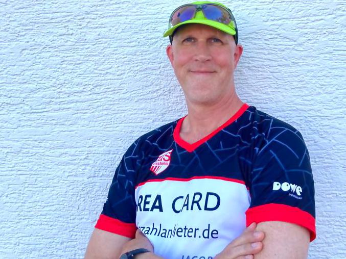 Mann mit Sonnenbrille und grünem Cap steht an einer weißen Wand, trägt ein T-Shirt mit Vereinslogo und gefalteten Armen.