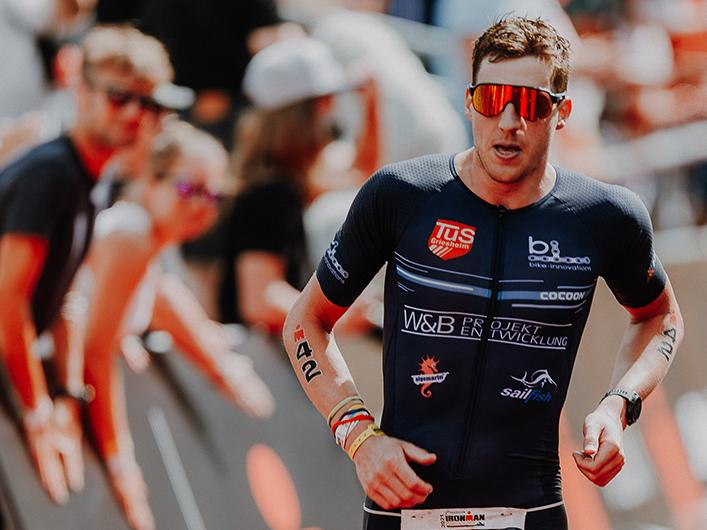 Sportler im Wettkampfanzug läuft konzentriert durch die Zuschauer, trägt die Nummer 42 und eine Sonnenbrille.