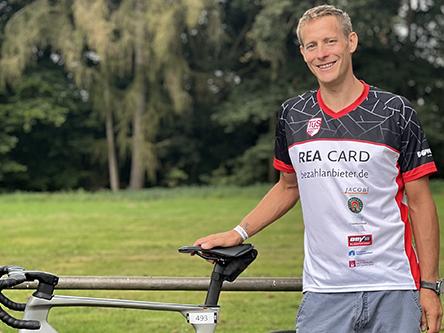 Ein Mann in einem Sporttrikot steht lächelnd neben einem Fahrrad auf einer grünen Wiese.