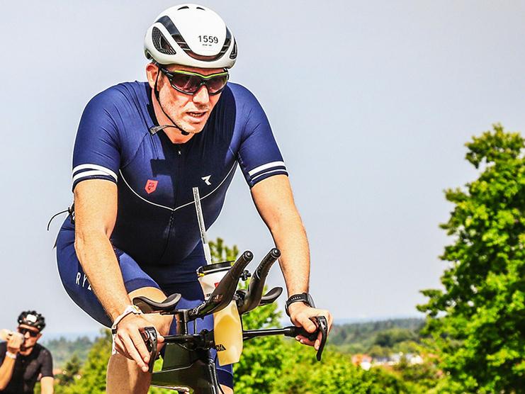 Radsportler in blauem Anzug fährt auf einer Straße, umgeben von anderen Radfahrern und grüner Landschaft.