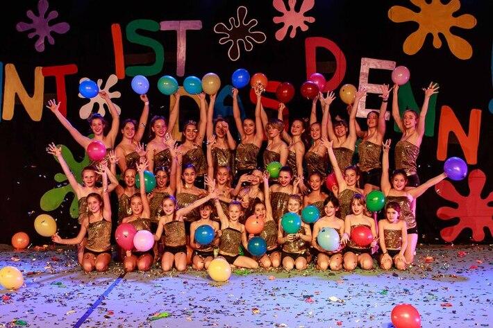 Gruppenaufführung mit Tänzerinnen in goldenen Kostümen, die bunte Luftballons vor einem farbenfrohen Hintergrund halten.