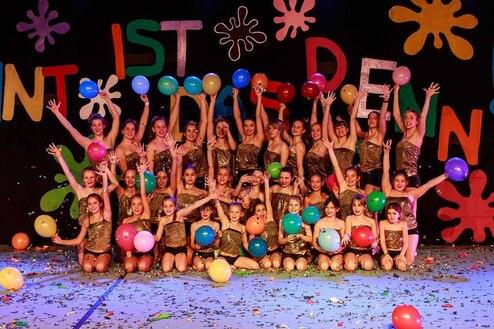 Gruppenaufführung mit Tänzerinnen in goldenen Kostümen, die bunte Luftballons vor einem farbenfrohen Hintergrund halten.