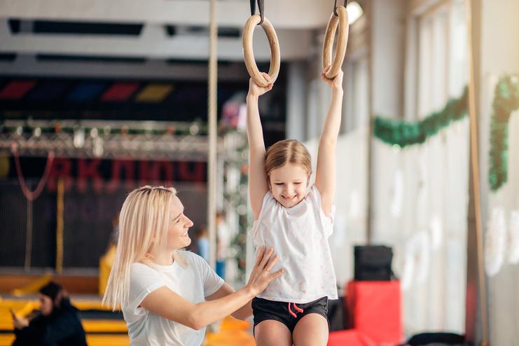Mädchen schwingt an Turnringen, während eine Trainerin sie lachend unterstützt und anlächelt. Sporthalle im Hintergrund.