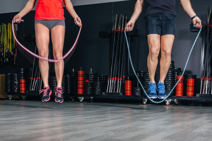 Zwei Personen springen mit Sprungseilen in einem Fitnessstudio auf einem Holzfußboden. Gewichte sind im Hintergrund sichtbar.