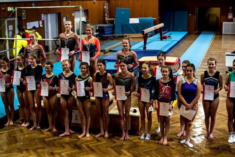 Gruppe junger Turnerinnen in Wettkampfanzügen hält Urkunden und Medaillen in einer Turnhalle.