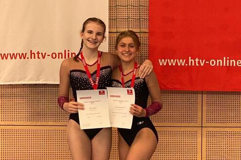 Zwei Sportlerinnen in Wettkampfkleidung halten Urkunden und l&auml;cheln vor einer Wand mit Banner.