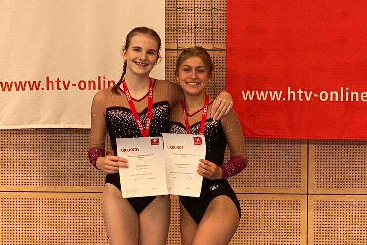 Zwei Sportlerinnen in Wettkampfkleidung halten Urkunden und lächeln vor einer Wand mit Banner.