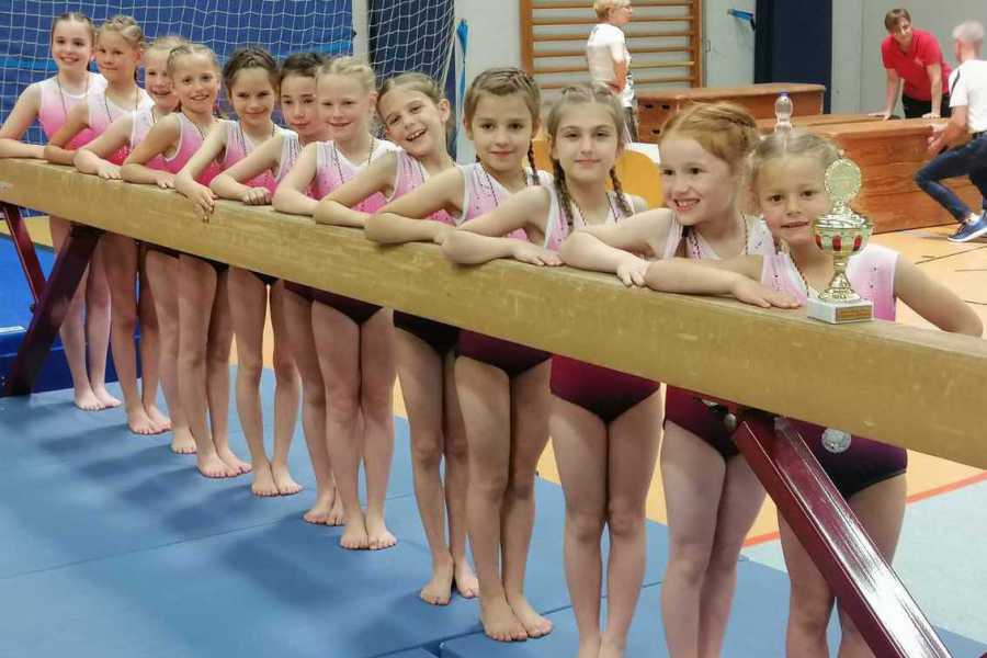 Gruppe junger Turnerinnen in Sportanzügen posiert an einem Barren mit einem Pokal auf einem Gymnastikboden.