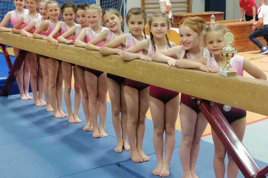 Gruppe junger Turnerinnen in Sportanzügen posiert an einem Barren mit einem Pokal auf einem Gymnastikboden.