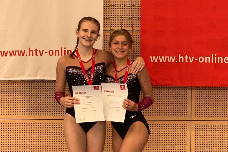 Zwei Sportlerinnen in Wettkampfkleidung halten Urkunden und lächeln vor einer Wand mit Banner.