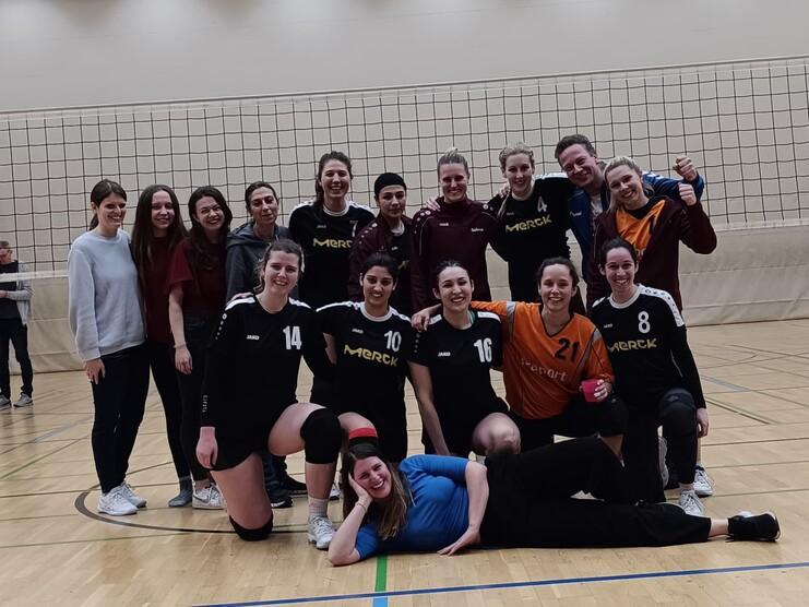 Gruppenselfie von Volleyballspielerinnen in Sportkleidung, mit Netz im Hintergrund und fröhlichen Gesichtern.