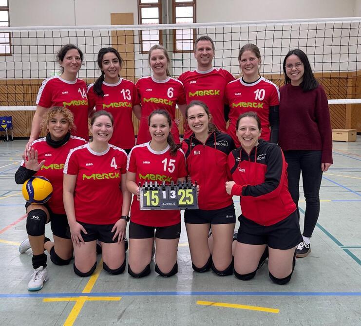 Gruppenfoto eines Volleyballteams in roten Trikots, mit einem Punktestand von 15:25 auf dem Schild in der Mitte.