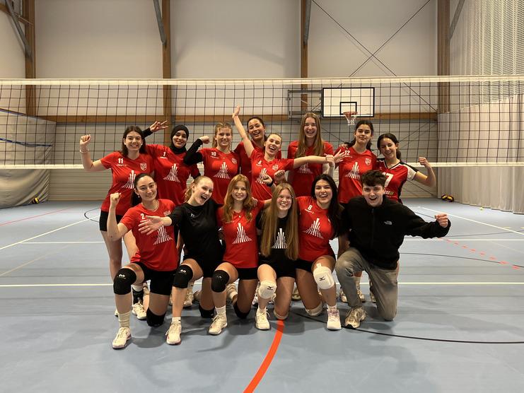 Gruppennfoto eines Volleyballteams in roten Trikots, fröhlich posierend auf einem Hallenboden mit Netz im Hintergrund.