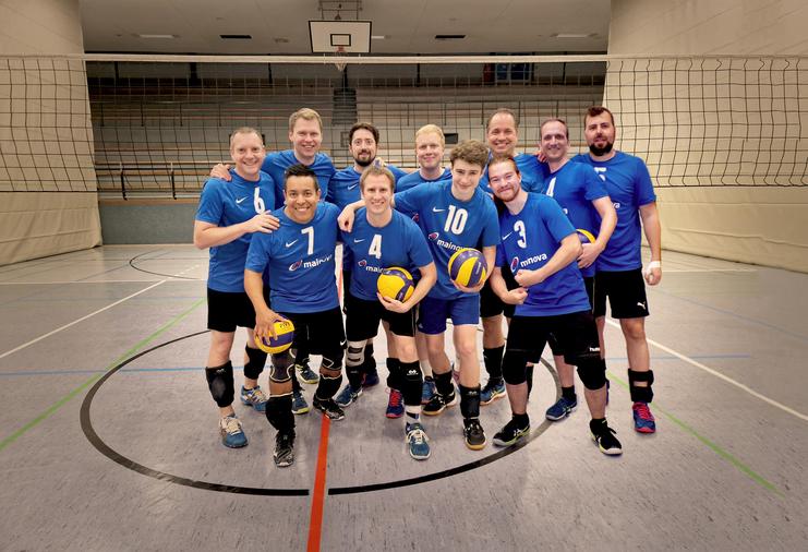 Gruppe von zehn Volleyballspielern in blauen Trikots mit Bällen, posierend auf einem Sportfeld hinter einem Netz.