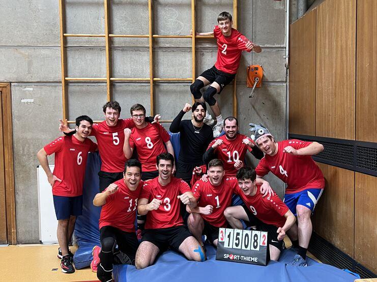 Gruppenselfie von zehn Männern in roten Trikots, die jubelnd auf einer blauen Matte stehen, mit einer Score-Anzeige im Vordergrund.