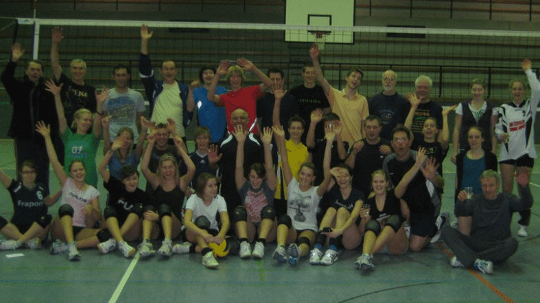 Gruppenevent mit etwa 30 Personen in Sportkleidung, die vor einem Volleyballnetz im Hallenboden posieren und jubeln.