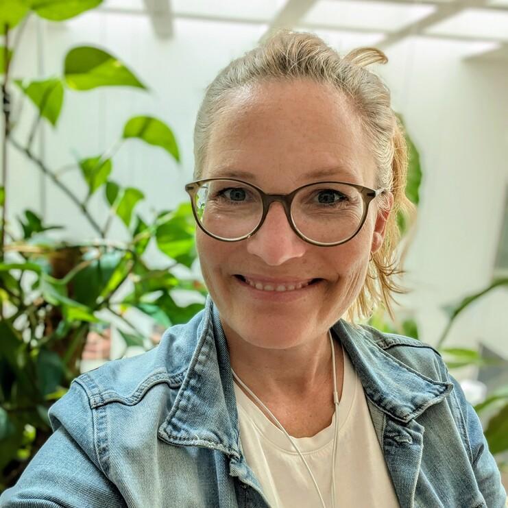 Frau mit Brille und Locken lächelt vor einer Wand aus grünen Pflanzen, trägt eine Jeansjacke und ein weißes T-Shirt.