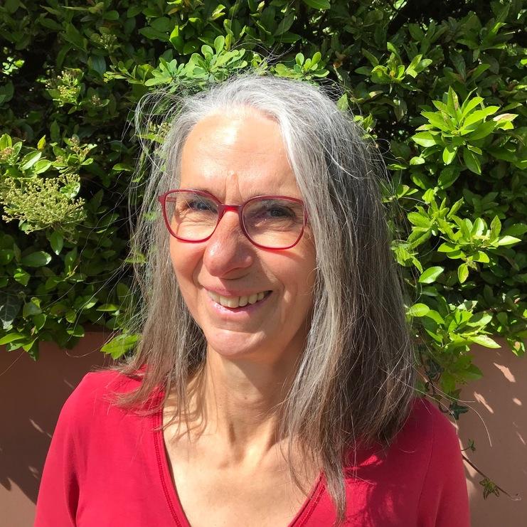 Frau mit grauen Haaren und roten Brillengestell lächelt vor grünem Hintergrund aus Büsche. Sie trägt ein rotes Oberteil.