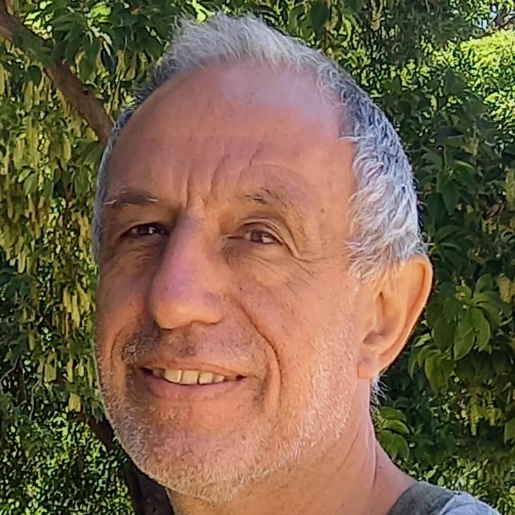 Mann mit grauen Haaren und kurzem Bart lächelt vor einem grünen Hintergrund aus Bäumen.