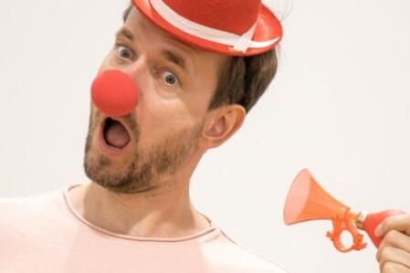 Mann mit roter Clownsnase und buntem Mini-Hut hält eine kleine rote Tröte und zeigt ein überrasches Gesicht.