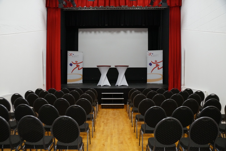 Veranstaltungsraum mit Stuhlreihen, zwei Stehtischen und Bühne hinter roten Vorhängen.