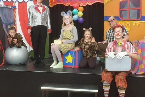 Sechs verkleidete Zirkuskünstler stehen auf einer Bühne mit buntem Hintergrund und Zirkusdekoration.