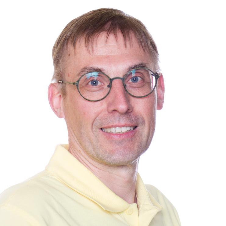 Porträt eines Mannes mit kurzen, glatten Haaren, Brille und gelbem Poloshirt vor weißem Hintergrund.