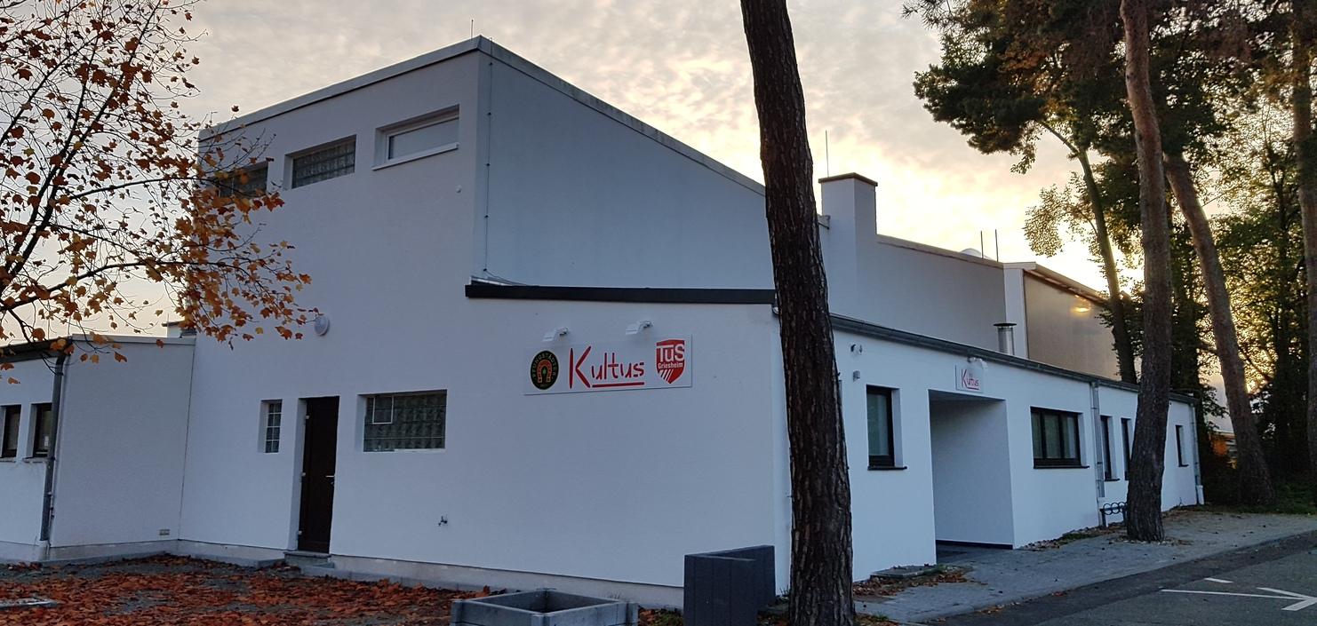 Modernes, zweigeschossiges Gebäude mit weißen Wänden, beschildert mit "Kultus", umgeben von herbstlichen Bäumen und Laub.