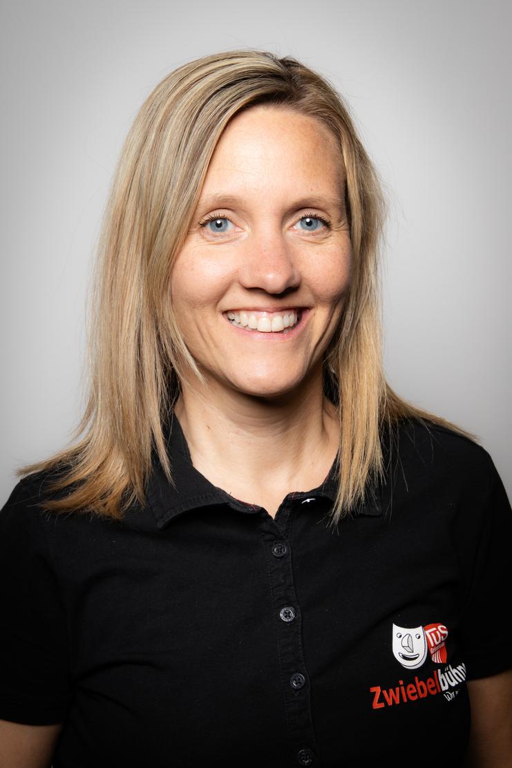 Porträt einer lächelnden Frau mit blonden Haaren, die ein schwarzes Polo mit Logo trägt, vor einem neutralen Hintergrund.