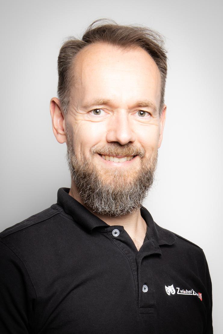 Porträt eines Mannes mit kurzem Haar und Vollbart, der ein schwarzes Polohemd trägt, lächelnd vor neutralem Hintergrund.