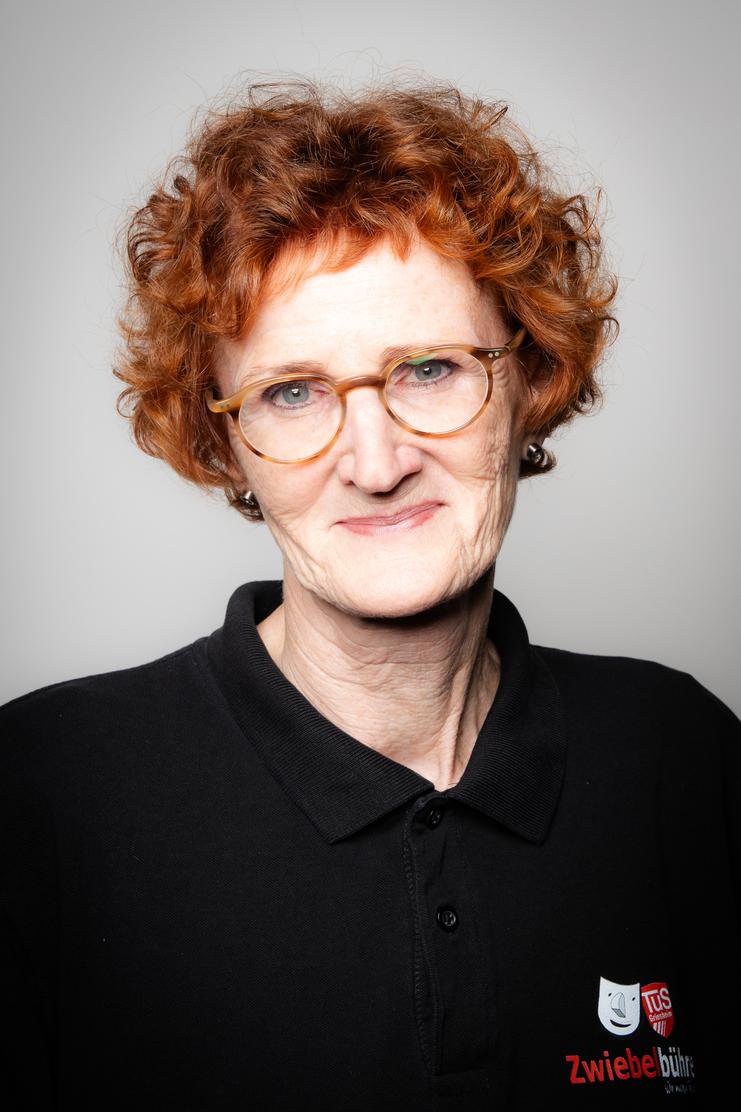 Porträt einer lächelnden Frau mit lockigem, rotem Haar, brilletragend, in schwarzem Poloshirt mit Logo.