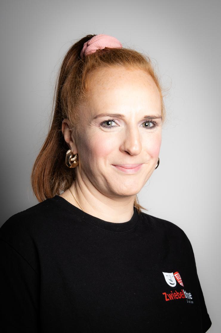 Porträt einer lächelnden Frau mit rotem Haar, einem rosa Haargummi und einem schwarzen T-Shirt mit Logo.