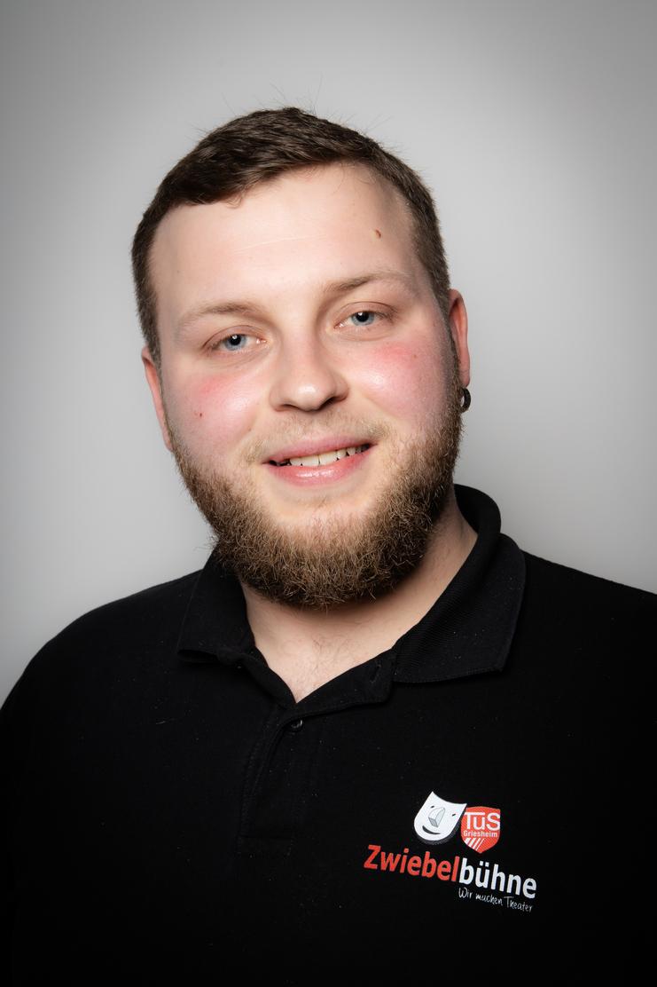 Porträt eines Mannes mit Vollbart, lächelnd, in schwarzem Poloshirt mit dem Logo der Zwiebelbühne. Hintergrund grau.