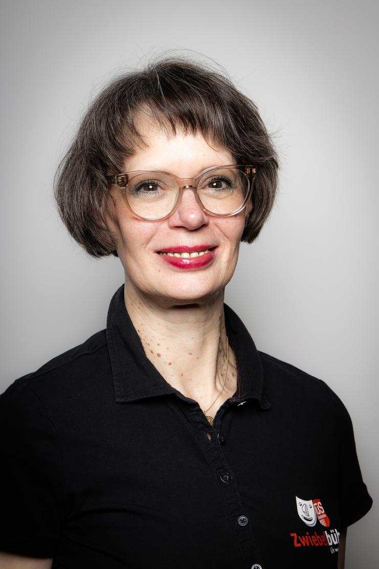 Porträt einer Frau mit kurzen, braunen Haaren und Brille, die ein schwarzes Hemd mit Aufdruck trägt. Hintergrund ist hell.