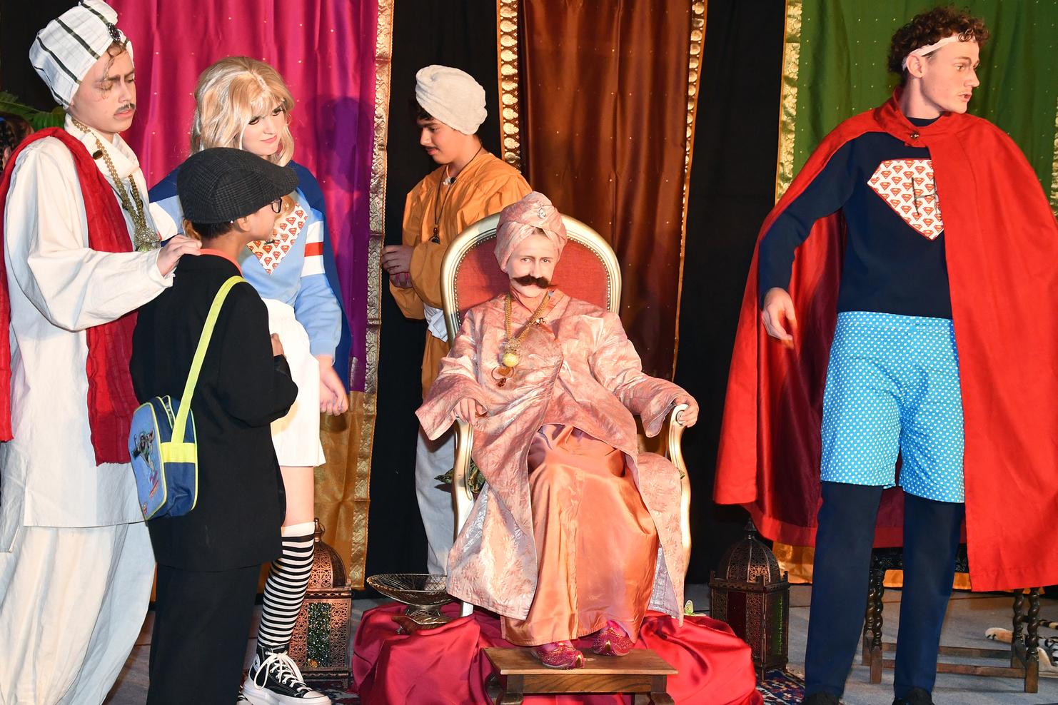 Szenenbild aus einem Theaterst&uuml;ck mit mehreren Kinderdarstellern in bunten Kost&uuml;men und einer sitzenden Figur in k&ouml;niglicher Kleidung.