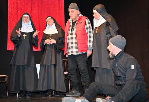 Vier Nonnen in schwarz-wei&szlig;en Gew&auml;ndern stehen um einen Mann in roter Weste auf einer B&uuml;hne mit schwarzem Hintergrund.