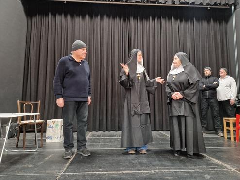 Drei Personen auf einer B&uuml;hne: zwei Nonnen im Habit stehen im Gespr&auml;ch, ein Mann beobachtet sie. Schwarzer B&uuml;hnenvorhang im Hintergrund.