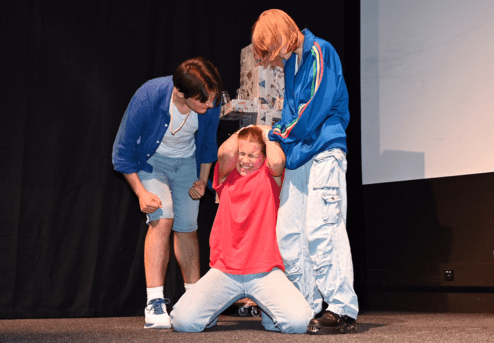 Drei Schauspieler auf einer Bühne: Zwei ziehen eine Person mit rotem Shirt, die schmerzverzerrt auf dem Boden kniet.