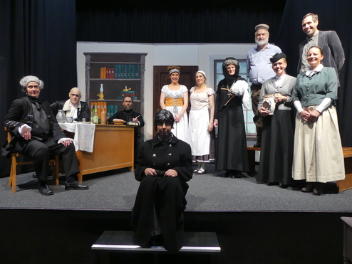 B&uuml;hnenbild mit zehn Schauspielern in historischen Kost&uuml;men auf einer Theaterb&uuml;hne, mit einem Tisch und Requisiten im Hintergrund.