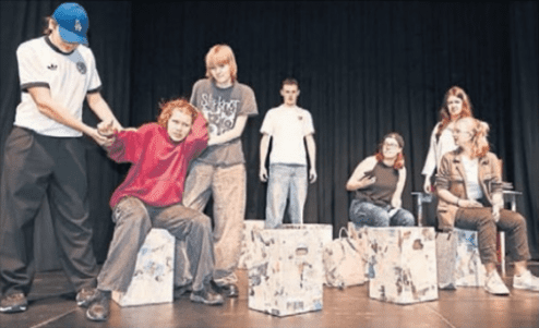 Acht Jugendliche in Kostümen auf einer Bühne, einige sitzen auf bemalten Kästen, andere interagieren miteinander.
