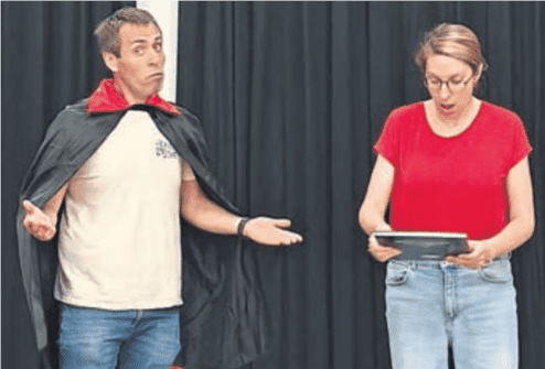 Ein Mann mit einem Umhang und einer lässigen Haltung steht neben einer Frau in einem roten Shirt, die ein Tablet hält.