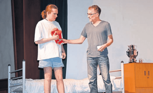 Zwei Schauspieler auf einer Bühne, einer in Pyjama mit einem Boxhandschuh, der andere überreicht etwas.