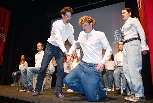 Schauspieler in weißen Hemden führen eine Szene auf einer Bühne mit von hinten sitzenden weiteren Darstellern auf.