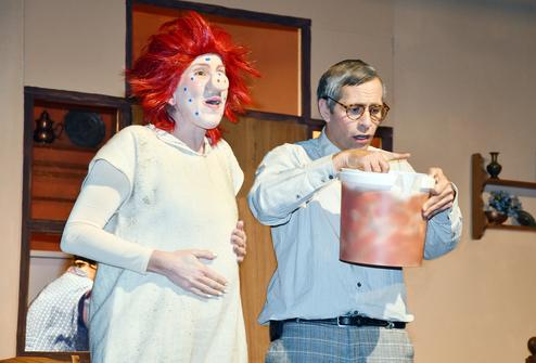 Zwei Schauspieler auf einer Bühne: ein clownesker Charakter und ein besorgter Mann mit einem Eimer in der Hand.