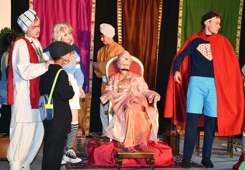 Szenenbild aus einem Theaterstück mit mehreren Kinderdarstellern in bunten Kostümen und einer sitzenden Figur in königlicher Kleidung.